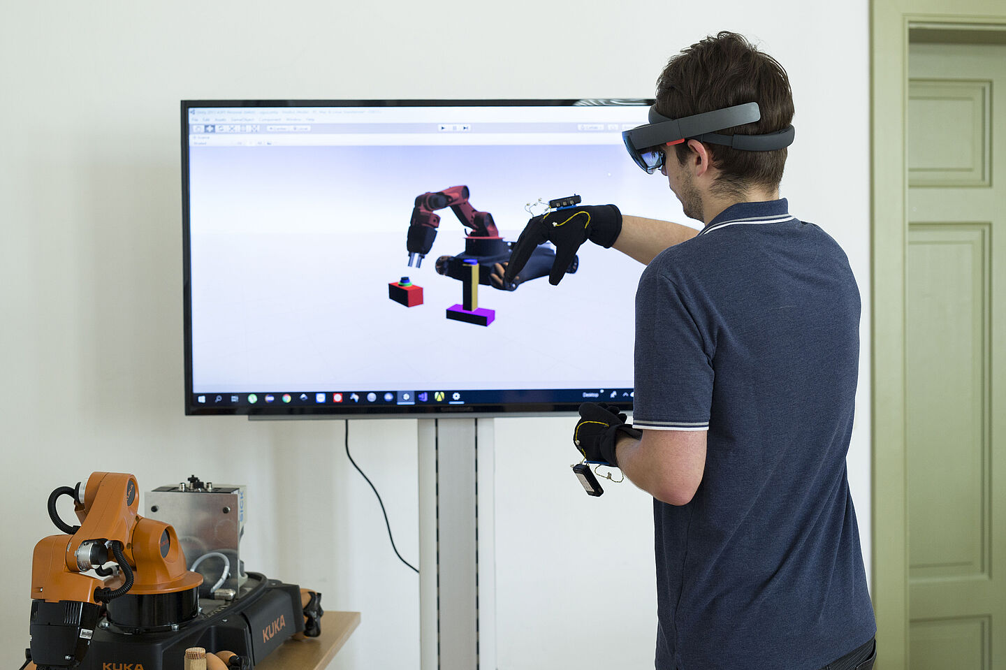 Mixed Reality - Roboter Steuerung vor Display Person mit VR-Brille und Joysticks bewegt in der virtuellen Realität Roboterteile. Das Ergebnis wird auf einem Bildschirm gezeigt.