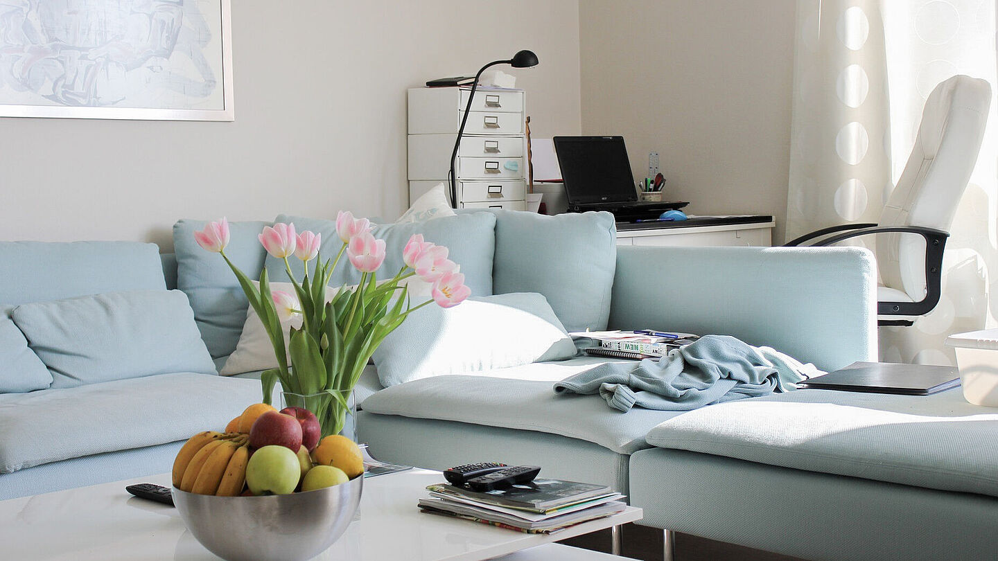 Wohnungen in Leipzig Ansicht helles Wohnzimmer mit Tisch, dahinter eine blaue Couch mit angrenzendem Arbeitsbereich